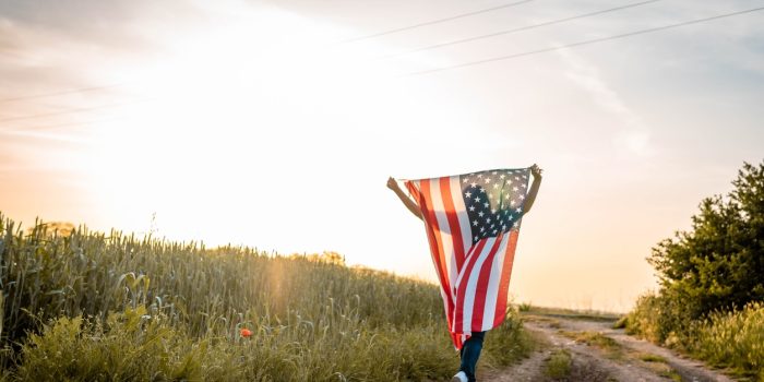 A Patriotic Woman Strolls A Rural Dirt Road Draping An American Flag Behind Her At Sunset