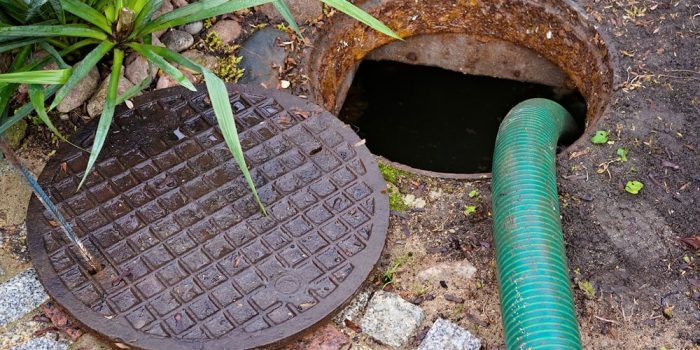 An Open Septic Tank In A Backyard With A Pump, Next To Some Plants And Rocks.