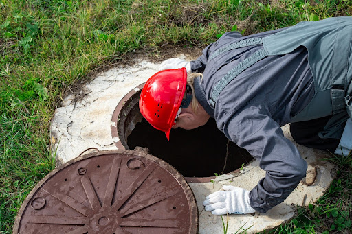 A Septic Technician With A Red Hard Har Peering Into A Septic Tank Through A Manhole.